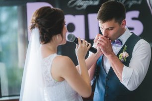 Жених в серой жилетке и фиолетовой бабочке целует руку невесты
