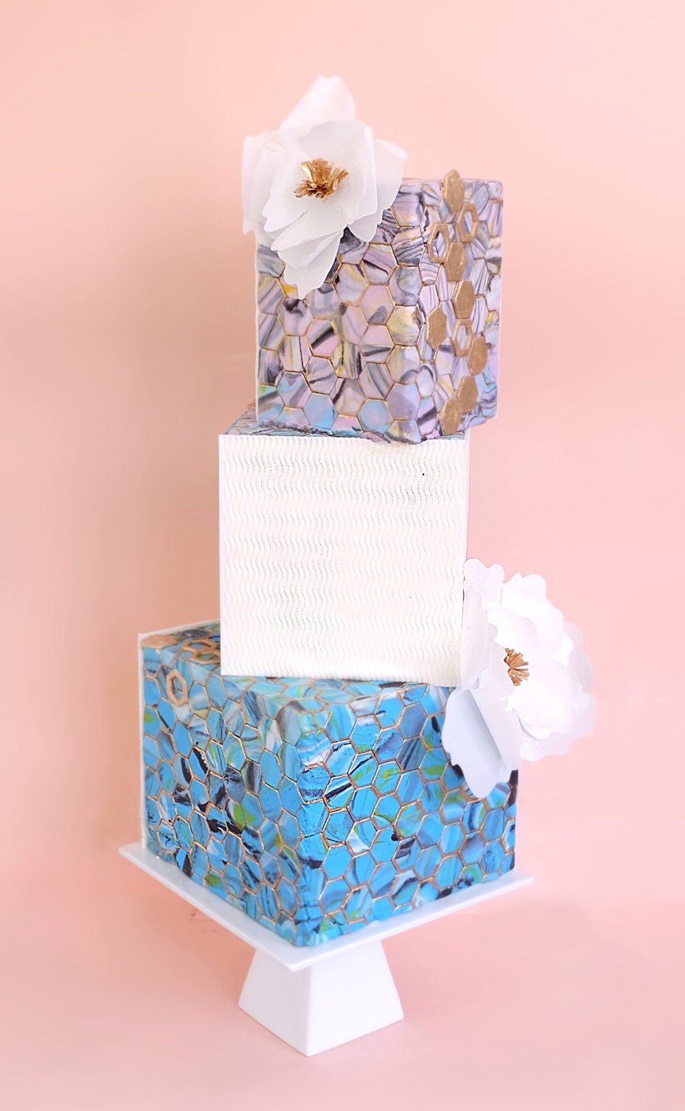 Стильный геометричный свадебный торт, покрытый мраморными шестигранниками из мастики, текстурированными белыми панелями и украшенный легкими вафельными цветами с золотыми серединками. 