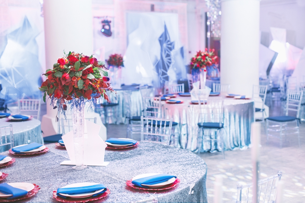 Оформление банкета в зимнем стиле. Периметр зала украшен белыми и серебряными декорациями в форме разнообразных снежных глыб, на столах серебряные пайеточные скатерти, прозрачные стулья, яркая флористика и сервировка