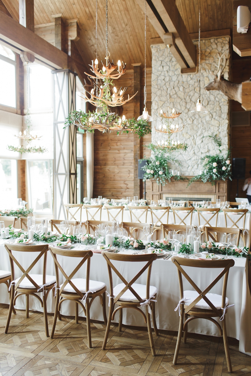 Столы гостей декорированы посередине гирляндой из зелени, цветов и свечей. Камин украшен свечами и композициями, деревянные стулья cross back. Люстры оформлены ветвями плетущейся зелени