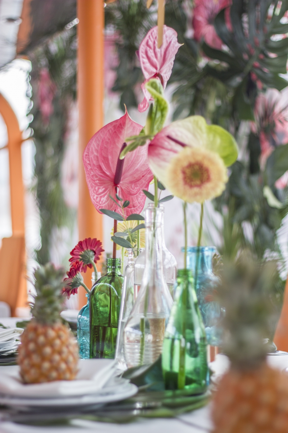 Тропический декор гостевых столов. Антуриум и герберы в разноцветных бутылочках, ананасики на тарелочках и листья монстеры под тарелками.