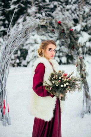 Невеста с букетом в платье цвета марсала на фоне свадебной арки зимой
